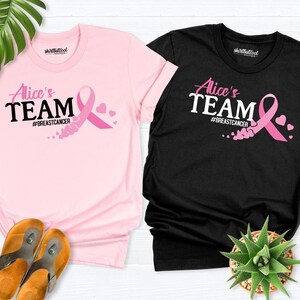 May include: Two t-shirts with the text "Alice's Team #BreastCancer" and a pink ribbon with hearts. The shirts are pink and black.