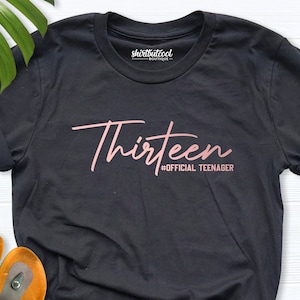 May include: Black t-shirt with the text "Thirteen #OFFICIAL TEENAGER" in pink lettering.