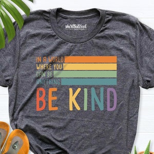 May include: A dark grey t-shirt with a rainbow stripe design and the text "In a world where you can be anything, be kind".