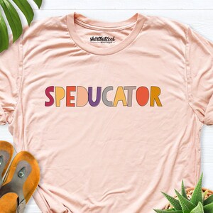 May include: A pink t-shirt with the text "SPEDUCATOR" in colorful letters. The letters are in different shades of red, orange, yellow, green, blue, purple, and gray.