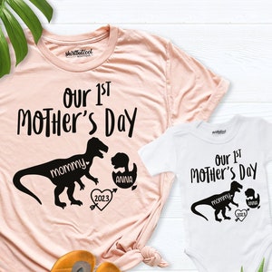 Peut inclure: Un t-shirt rose et un body blanc avec un motif de dinosaure et le texte "Our 1st Mother's Day". Le dinosaure sur le t-shirt dit "mommy" et le dinosaure sur le body dit "mommy". Le dinosaure sur le body a un plus petit dinosaure dans sa bouche qui dit "anna". Les deux t-shirts ont un c&oelig;ur avec l'ann&eacute;e 2023.