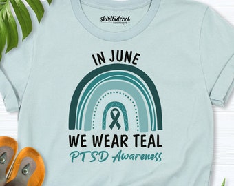 以下が含まれることがあります： 薄い青色のTシャツに、ターコイズブルーの虹とリボンがデザインされています。Tシャツには「In June We Wear Teal PTSD Awareness」という文字が印刷されています。