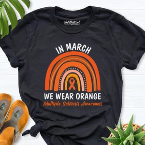 May include: Black t-shirt with the text "IN MARCH WE WEAR ORANGE Multiple Sclerosis Awareness" above a rainbow design in shades of orange. The shirt is styled with a knot at the bottom. A pair of orange sandals and a succulent plant are also in the image.