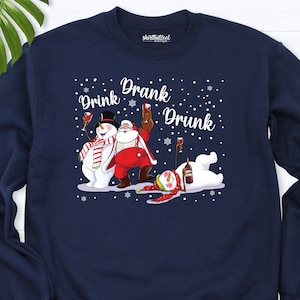 May include: Navy blue sweatshirt with a festive graphic featuring a snowman, Santa Claus, and a snowman, all holding wine glasses. The text "Drink Drank Drunk" is displayed above the characters in a white font. Snowflakes are scattered around the characters.