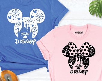 May include: Two t-shirts with a Disney theme. The blue shirt has a white silhouette of Mickey Mouse's head with fireworks and Cinderella's castle inside. The pink shirt has a black silhouette of Minnie Mouse's head with a polka dot bow and fireworks and Cinderella's castle inside. Both shirts have the text "2025 DISNEY" underneath the silhouette.