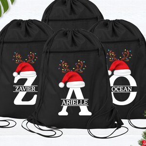 May include: Four black drawstring bags with a Christmas theme. Each bag features a red Santa hat, reindeer antlers, and a letter with a name below it. The names are Zavier, Arielle, and Ocean.