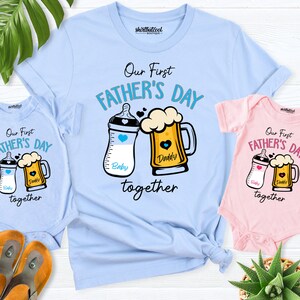 May include: A set of three light blue and pink t-shirts with the text "Our First Father's Day together" and a graphic of a baby bottle and a beer mug. The baby bottle is labeled "Baby" and the beer mug is labeled "Daddy".