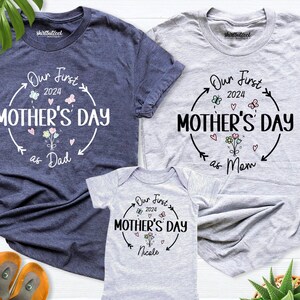 May include: Three grey t-shirts with a white graphic design that says "Our First Mother's Day" with a flower and heart design. The shirts are for a family of three, with the largest shirt saying "as Dad", the medium shirt saying "as Mum", and the smallest shirt saying "Nicole".
