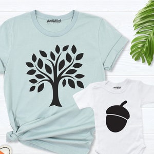 May include: A light blue t-shirt with a black tree design and a white bodysuit with a black acorn design. The shirts are on a white wooden background.