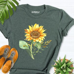 May include: A dark green t-shirt with a watercolor sunflower design. The sunflower is yellow with a brown center and green leaves. The shirt has a rolled up sleeve and is laid flat on a white wooden surface.