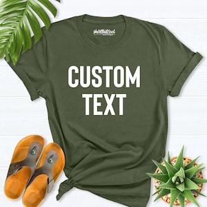 May include: A green t-shirt with the text "CUSTOM TEXT" printed in white on the front. The shirt is laid flat on a white wooden surface.