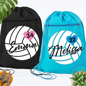 May include: Two drawstring backpacks, one black and one turquoise. Each features a white volleyball design with a name and number in a heart. The black bag has "Emma" and "34" in a pink heart. The turquoise bag has "Melissa" and "22" in a blue heart.