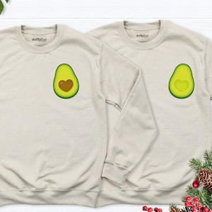 May include: Two beige sweatshirts with a green avocado design featuring a brown heart in the center. The text "shortfuze" is printed on the collar of each sweatshirt.