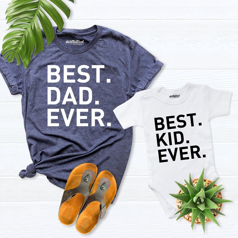May include: Two t-shirts, one blue and one white, with the text "Best Dad Ever." and "Best Kid Ever." printed on them. The shirts are laid out on a white wooden surface with a pair of brown sandals.