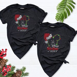 Peut inclure: Deux t-shirts noirs avec un motif de t&ecirc;te de Mickey Mouse scintillant avec un chapeau de P&egrave;re No&euml;l et les mots "Disney Christmas 2023".