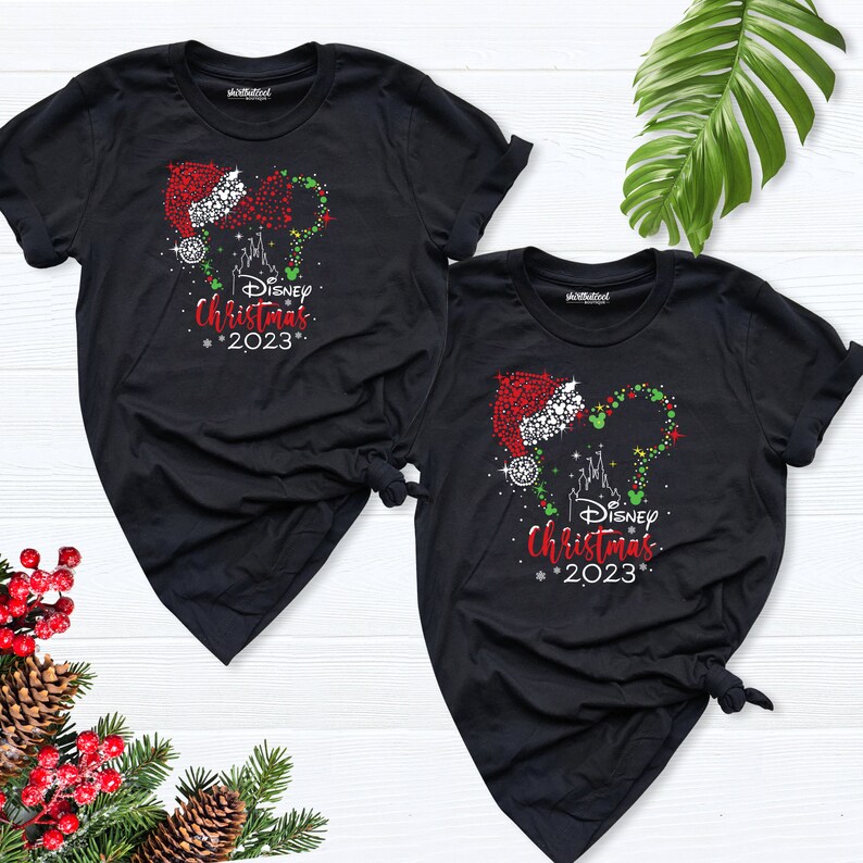 Peut inclure: Deux t-shirts noirs avec un motif de t&ecirc;te de Mickey Mouse scintillant avec un chapeau de P&egrave;re No&euml;l et les mots "Disney Christmas 2023".