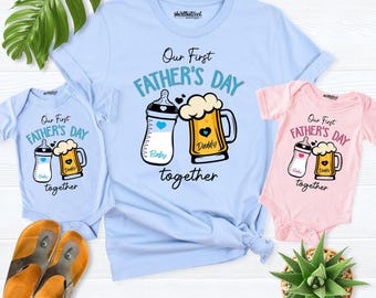 May include: A light blue adult t-shirt and two baby onesies, one light blue and one pink. Each garment features a graphic of a baby bottle and a beer mug with the text Our First FATHER'S DAY together. The baby bottle is labeled Baby and the beer mug is labeled Daddy.