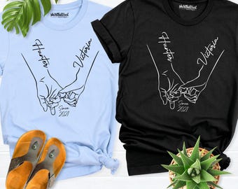 以下が含まれることがあります： ライトブルーのTシャツとブラックのTシャツ。どちらも小指をつないだ2本の手の白い線画が描かれています。手の上にはHunterとVictoriaの文字があり、指の下にはSince 2021の文字がプリントされています。