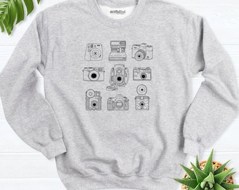 May include: A light gray crewneck sweatshirt featuring a black line art graphic of nine different vintage cameras arranged in a grid. The interior neck label displays the text skirtbuttool boutique in a simple black font.
