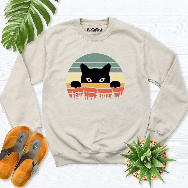 K&ouml;nnte beinhalten: Ein beiger Pullover mit einem Retro-Grafikdruck eines schwarzen Katers, der &uuml;ber einen Sonnenuntergang mit B&auml;umen schaut. Der Text "shirtbutcool" ist auf dem Pullover gedruckt.