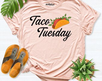 May include: A light pink short-sleeve t-shirt featuring the black text Taco Tuesday and a graphic of a taco. The shirt has a crew neckline and a label inside the collar that reads shirtbutcool BOUTIQUE.