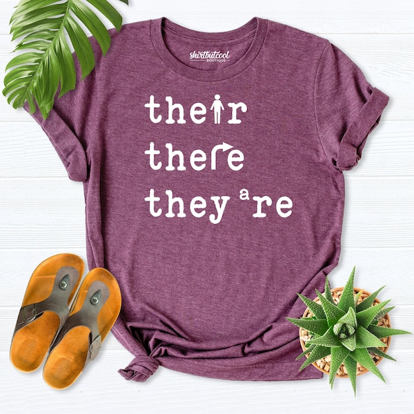 May include: A heathered maroon short-sleeve t-shirt featuring white text that reads their, there, and they're. The letter i in their is a stick figure, the r in there is an arrow, and the a in they're is a small letter a.