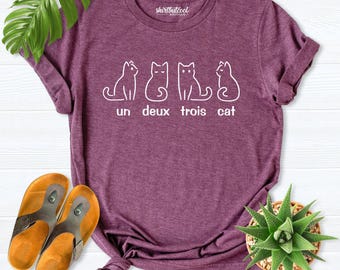 Può includere: Una t-shirt a maniche corte color bordeaux mélange con una grafica a linee bianche di quattro gatti sopra la scritta "un deux trois cat". La maglia presenta un girocollo ed è realizzata in un tessuto morbido e testurizzato con etichetta.