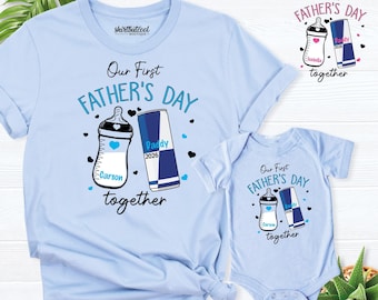 May include: A light blue t-shirt and a matching baby bodysuit featuring a graphic of a baby bottle and an energy drink can. The text reads Our First Father's Day together, with the names Carson and Daddy, and the year 2026 printed on the items.