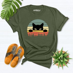 K&ouml;nnte beinhalten: Ein gr&uuml;nes T-Shirt mit einer schwarzen Katze, die &uuml;ber einem Retro-Sonnenuntergang-Design lugt. Der Text "shirtbutcool - boutique" ist auf das T-Shirt gedruckt.