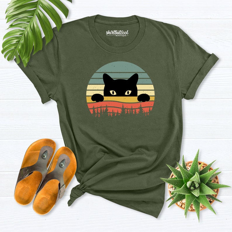 K&ouml;nnte beinhalten: Ein gr&uuml;nes T-Shirt mit einer schwarzen Katze, die &uuml;ber einem Retro-Sonnenuntergang-Design lugt. Der Text "shirtbutcool - boutique" ist auf das T-Shirt gedruckt.