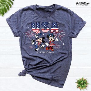 Pode incluir: Uma t-shirt azul-marinho mesclado com um gr&aacute;fico do Mickey e da Minnie vestidos com trajes patri&oacute;ticos. O design inclui o texto USA, fogo de artif&iacute;cio e os anos 1776-2026. A t-shirt tem mangas curtas e um decote redondo.