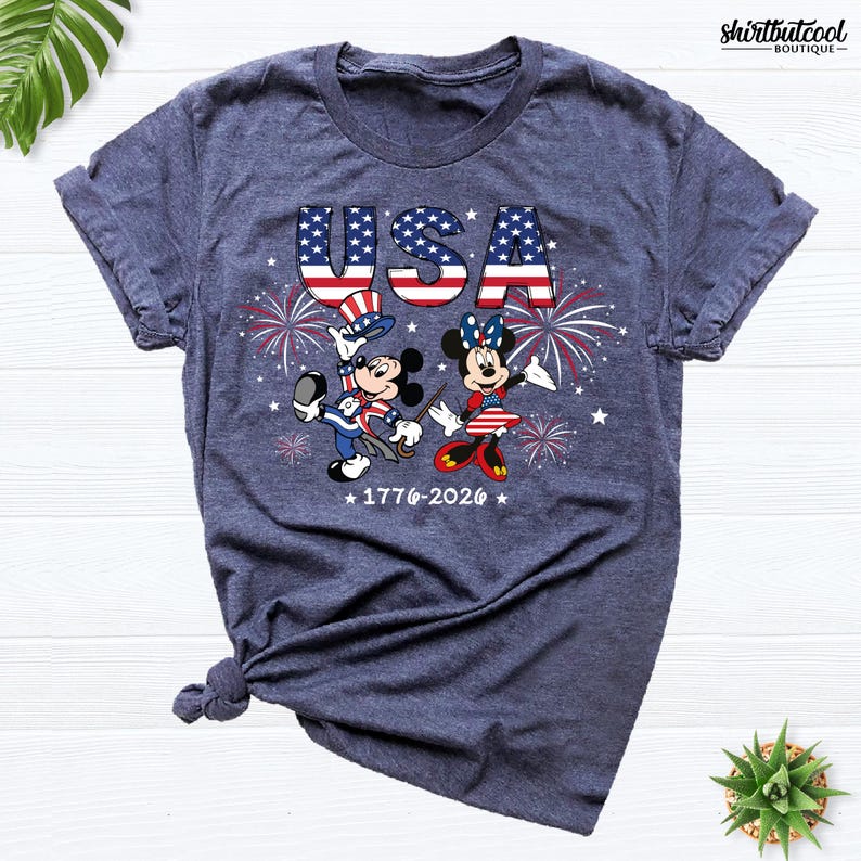 Pode incluir: Uma t-shirt azul-marinho mesclado com um gr&aacute;fico do Mickey e da Minnie vestidos com trajes patri&oacute;ticos. O design inclui o texto USA, fogo de artif&iacute;cio e os anos 1776-2026. A t-shirt tem mangas curtas e um decote redondo.