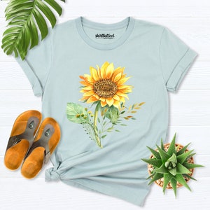 May include: A light blue t-shirt with a watercolor sunflower design. The sunflower is yellow with a brown center and green leaves. The shirt has the text "shirtbutcool" printed on the front.
