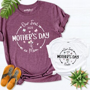 May include: Two t-shirts with a floral design and the text "Our First Mother's Day" on them. The larger shirt is purple and says "as Mom" below the text. The smaller shirt is white and says "Nicole" below the text.