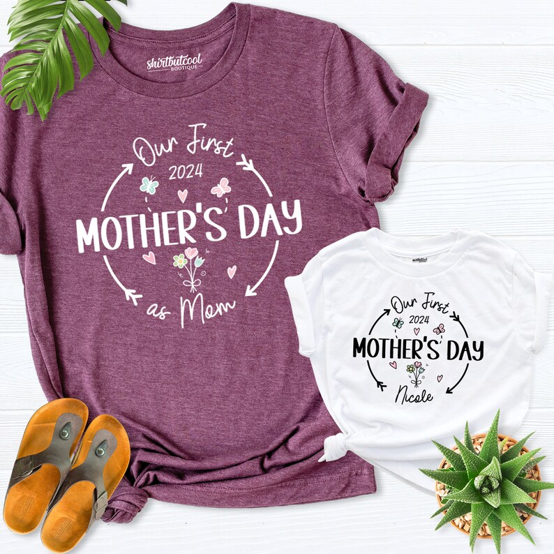May include: Two t-shirts with a floral design and the text "Our First Mother's Day" on them. The larger shirt is purple and says "as Mom" below the text. The smaller shirt is white and says "Nicole" below the text.