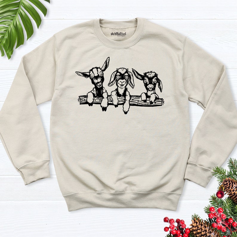 K&ouml;nnte beinhalten: Ein beiger Pullover mit einer schwarzen Grafik von drei jungen Ziegen, die &uuml;ber einen Ast schauen. Die Ziegen haben schwarze Augen und Nasen und schauen den Betrachter an.