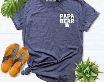 May include: A heather navy blue short-sleeve t-shirt featuring the white text PAPA BEAR above a bear paw print graphic on the left chest area. The shirt includes a small white logo at the inner neckline that reads shirtbutcool boutique.