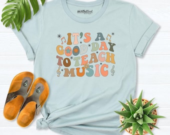 Może przedstawiać: Jasnoniebieski t-shirt z krótkim rękawem z napisem "IT'S A GOOD DAY TO TEACH MUSIC" wykonanym wielokolorową, zaokrągloną czcionką. Wzór zawiera małe nuty, klucze wiolinowe oraz akcenty w kształcie gwiazdek w kolorze pomarańczowym, morskim i brązowym.