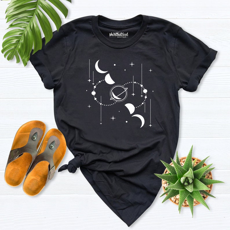 May include: Black t-shirt with a white graphic design of a celestial scene with moons, stars, and a planet. The text "shirtthatcool" is printed on the shirt.