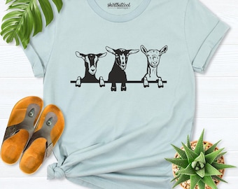 May include: A light blue t-shirt featuring a black graphic of three goats peeking over a horizontal line. The shirt includes the text "shirtbutcool BOUTIQUE" printed in black on the inside neck area. The design is centered on the chest.