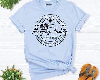 May include: A light blue t-shirt featuring a black circular graphic with palm trees, a sun, birds, and the text "FAMILY VACATION", "Murphy Family", "Florida 2025", and "MAKING MEMORIES TOGETHER" printed on the front center.