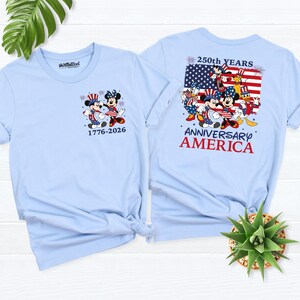 May include: Light blue t-shirts featuring Mickey and Minnie Mouse in patriotic attire. One shirt displays "1776-2026" and the other has "250th YEARS ANNIVERSARY AMERICA" with an American flag design. The shirts are a celebration of American history.