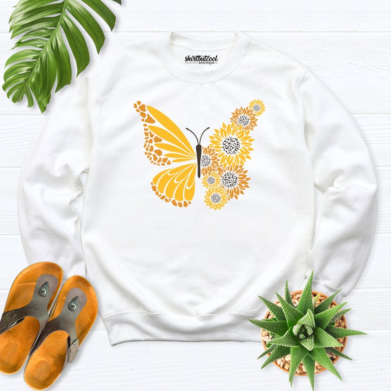 May include: White sweatshirt with a yellow butterfly design made of sunflowers. The text "shirtbutcool" is printed on the front of the sweatshirt.