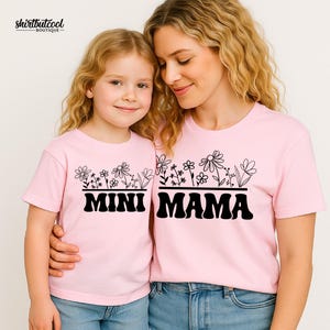 Pode incluir: Duas camisetas de manga curta rosa claro com um gráfico preto de flores silvestres acima do texto MINI em uma camiseta e MAMA na outra. As camisetas têm gola redonda e são combinadas com jeans azuis. O logotipo SHIRTBUTCOOL BOUTIQUE é visível.