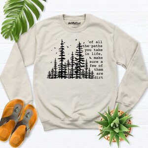 May include: A beige sweatshirt with a black forest silhouette and the text 'of all the paths you take in life, make sure a few of them are dirt'.
