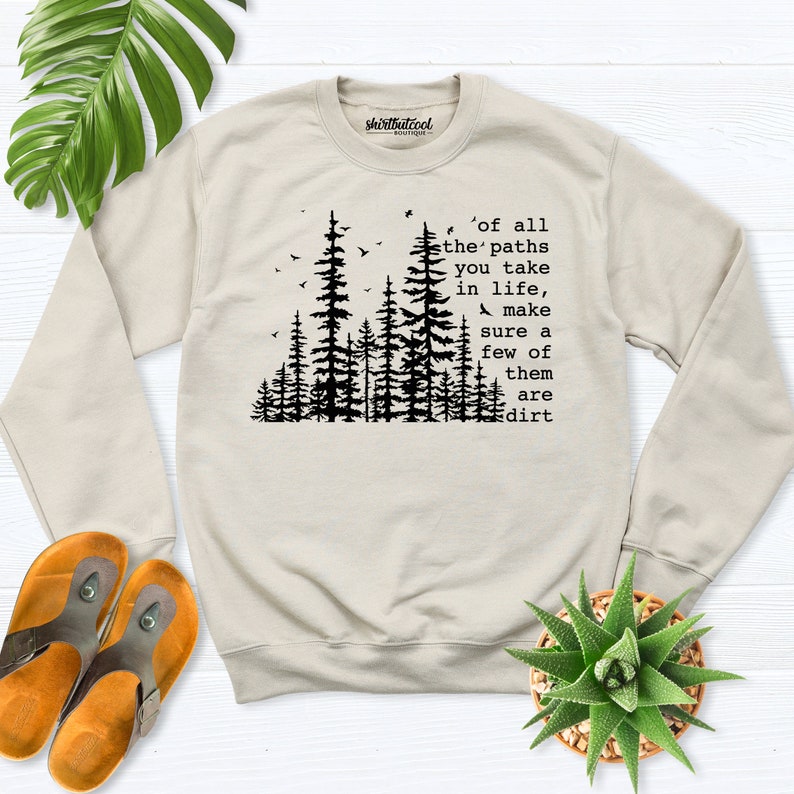 May include: A beige sweatshirt with a black forest silhouette and the text 'of all the paths you take in life, make sure a few of them are dirt'.