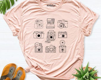 May include: A light pink short-sleeve t-shirt featuring a black line art graphic of nine different vintage cameras arranged in a grid. The inner neck label displays the text shirtbutcool boutique in black lettering.