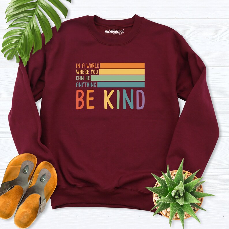 May include: Burgundy sweatshirt with a retro-style graphic that reads "In a world where you can be anything, be kind". The graphic features colorful stripes.