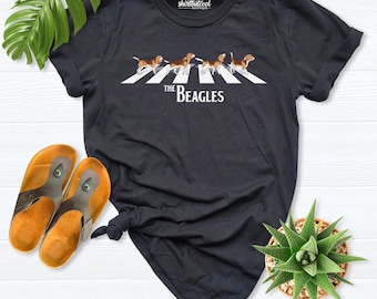May include: A black short-sleeved t-shirt featuring a graphic of four beagle dogs walking across a white crosswalk, with the text "THE BEAGLES" printed below the dogs. The shirt has a crew neckline and a label reading "shirtbutcool".