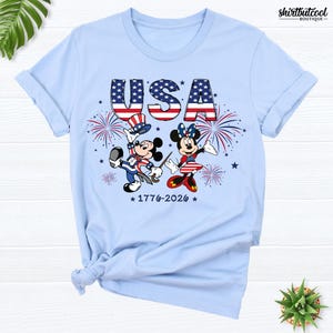 Pode incluir: Uma t-shirt azul-clara com um gr&aacute;fico do Mickey e da Minnie vestidos com trajes patri&oacute;ticos. Acima, o texto USA est&aacute; impresso com um padr&atilde;o de estrelas e riscas. Fogo de artif&iacute;cio e o texto 1776-2026 tamb&eacute;m aparecem na frente.